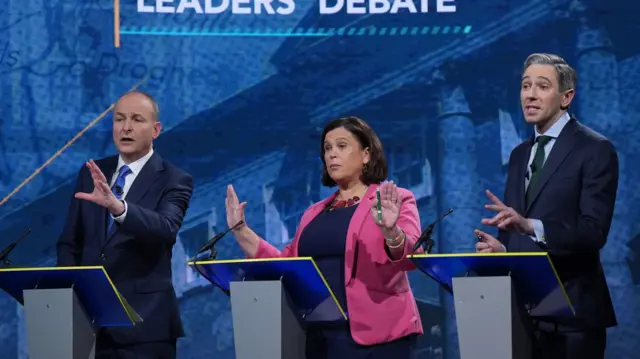 Micheal Martin wears a navy suit and blue tie standing behind a plinth. Mary Lou McDonald is doing the same, she wears a pink blazer and Navy dress. Simon Harris, on the far right, has on a green tie and blue shirt with a navy blazerr