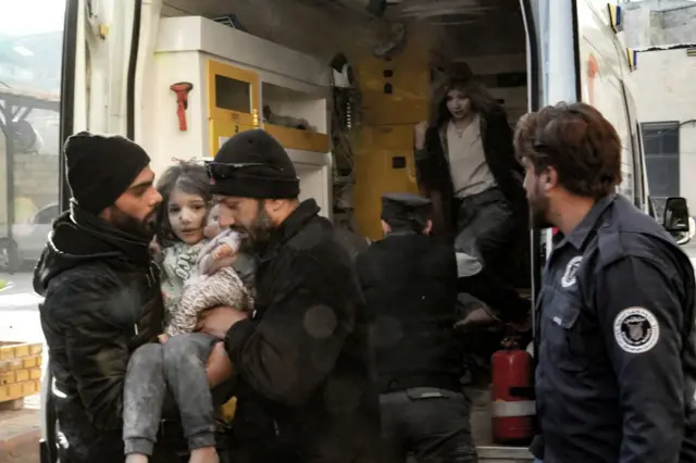 An injured girl and a woman in an ambulance. They're both grey from ash, the girl is being lifted by men out of the ambulance as she holds a doll