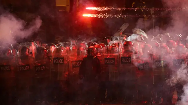 Fireworks can be seen just a little above the heads of scores of riot police