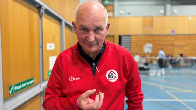 A man in a red quarter zip hold a gold ring behind him, out of focus, are people counting election ballots.