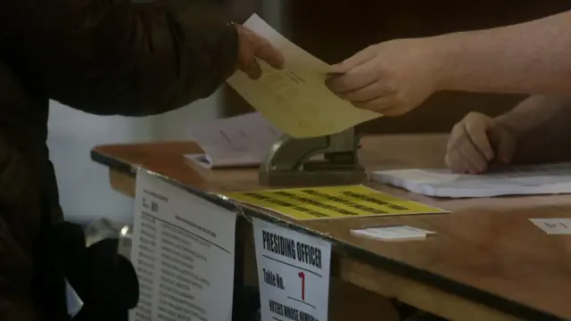 Two people exchange a ballot paper over a wooden desk