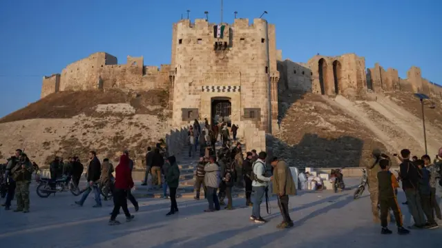 Syrian opposition fighters stand outside of a large citadel, which has a flag draped over the top of it.