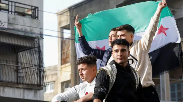 People celebrate in Syria, holding a green, white and black Syrian flag with buildings in the background