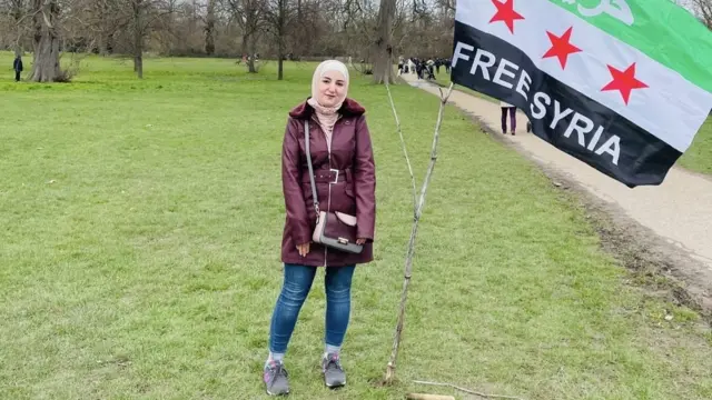 Douna Haj Ahmed stands outside in a purple jacket and blue jeans next to a flag hung on a branch reading Free Syria