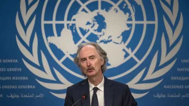 United Nations (UN) Special Envoy for Syria stands behind a podium and in front of a large backdrop image of the UN logo