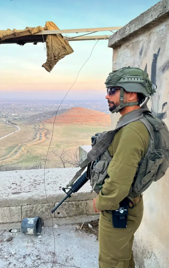 IDF solider in combat gear carrying an assalt rifle standing near the edge of a building, a distinctive hilltop can be seen in the background