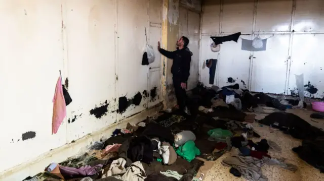 A man in a windowless room in the Saydnaya prison. There is clothing and debris littering the floor