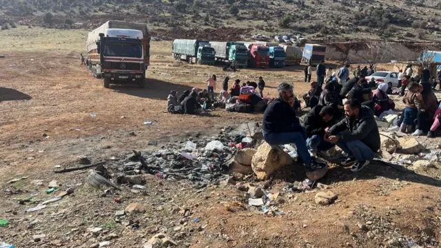 People sit near the border crossing, surrounded by rubble