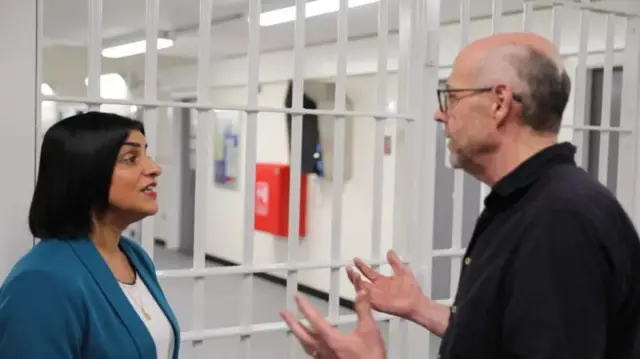 Shabana Mahmood in a blue blazer in front of a white prison gate speaking to Nick Robinson (on the right) in  a black shirt facing her with his hands raised up