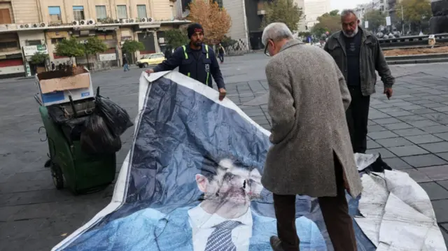 A man walks on a poster of Bashar al-Assad as a sanitation worker removes it from the street downtown