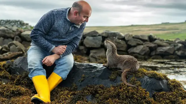 A man sits on a rock on the coast wearing yellow wellingtoon boots looking down at an otter sat on the same rock, looking up at the man