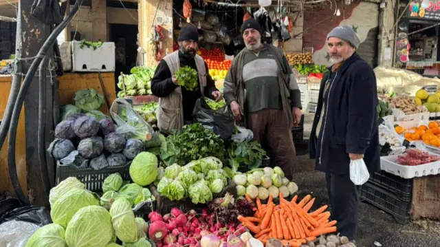 Three men in Damascus stand outside a vegetable market stall, one an is holding some green leaves in his hands while the other two men look towards the camera