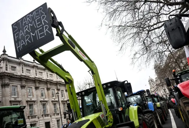 A convoy of tractors lines a central London road. The one in front holds up a sign that reads Starmer Farmer Harmer