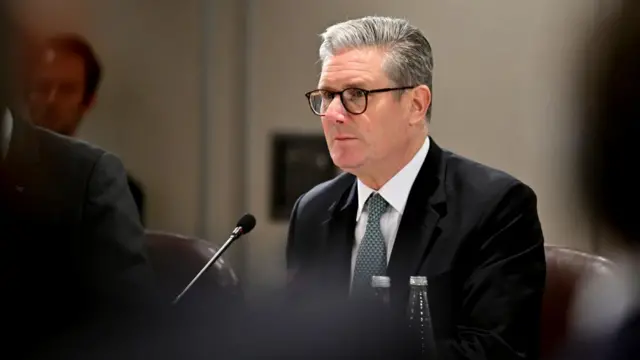 Starmer, wearing a suit, sits at a table with a small microphone in front of him