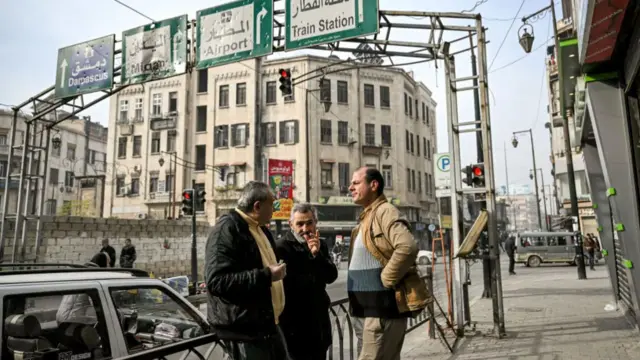 Three men chat on pavement under street signs for Damascus airport and train station. A bike is in the street at a red signal