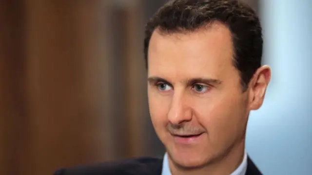 Bashar al-Assad close up with mouth partially open. He's sitting down wearing a dark suit and light blue shirt