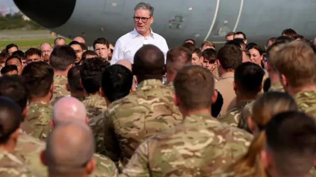 Starmer stands on a raised platform, just his head and shoulders can be seen over the top of a crowd of people in army fatigues standing around him