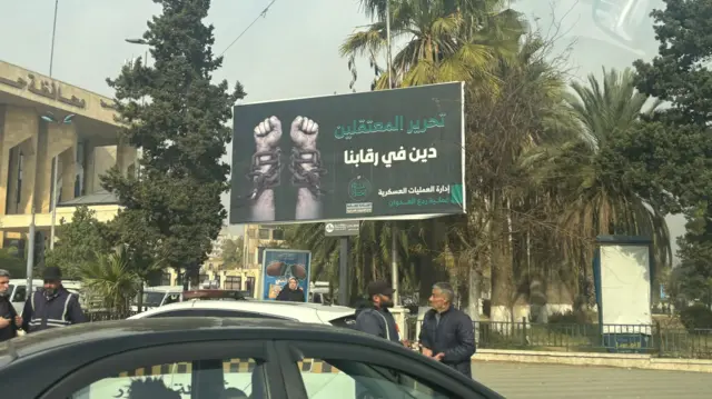 Four men stand in the streets, parked cars around them. In the background is a billboard in black showing two chained arms with raised fists reading "Freeing detainees is a debt upon our necks" in Arabic