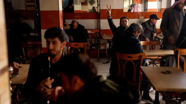 A man holds up a peace sign inside a Syrian coffee and shisha bar while men sit around him smoking shisha pipes