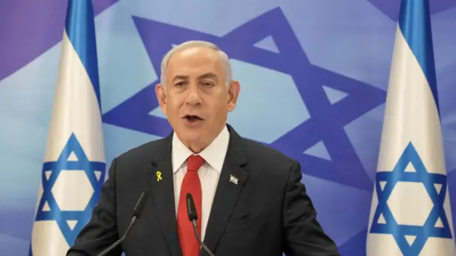 Benjamin Netanyahu delivers speech standing in black suit, white shirt and red tie. Behind him are two Israeli flags and a wall decorated with the flag, Star of David featuring prominently at the centre