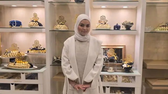 Damascus chocolate shop worker Joud stands in the centre of the photo in white coat and cream jumper, her hands together in front of her. She's wearing a white head scarf as she stands at the centre of the shop