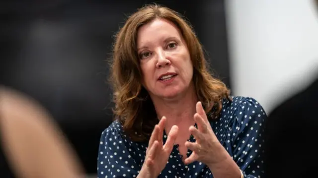 A picture of Souza talking whilst gesturing with her hands. She wears a polka dot top and a gold bracelet