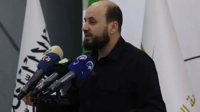 New interim Syrian prime minister Mohammed al-Bashir speaks to the press. He's standing in front of a lectern holding three mics wearing a black shirt. Behind him are two flags