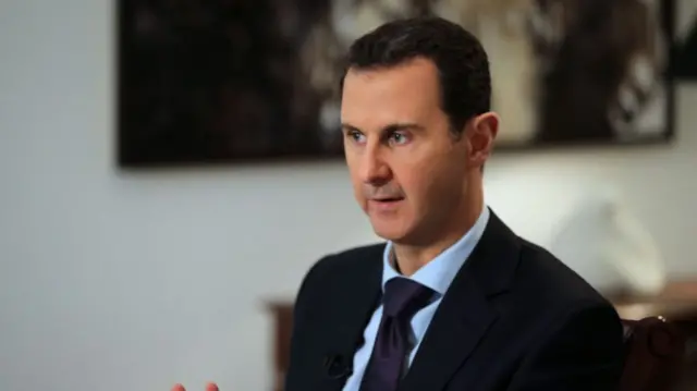 Bashar al-Assad sitting down mid-speech. He's wearing a black suit, deep blue tie and light blue shirt. A wooden table with a decorative white vase is visible behind him, above is a painting (blurred)