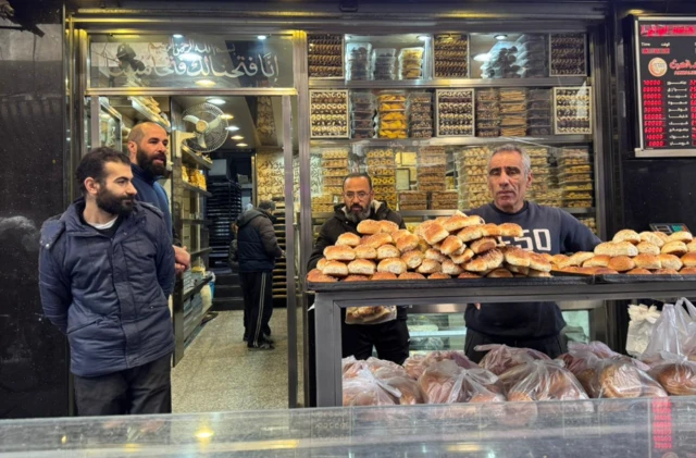 Three men stand outside a Syrian bakery in Damascus with loaves of bread piled up on a table outside the shop