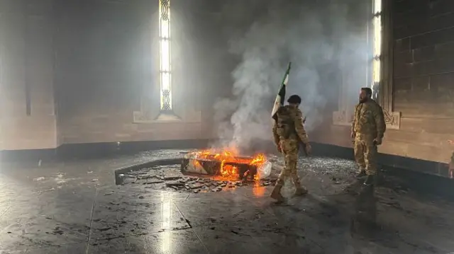 Rebel fighters stand next to the burning gravesite of Syria's late president Hafez al-Assad. What appears to be a wooden coffin is ablaze at the centre of the image, with two men in military fatigues standing to its left. The man in the foreground is carrying a Syrian opposition flag