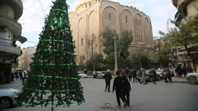 A Christmas tree in the centre of Aleppo, people walking down the streets. Residential buildings to the side and a religious building in stone at the centre