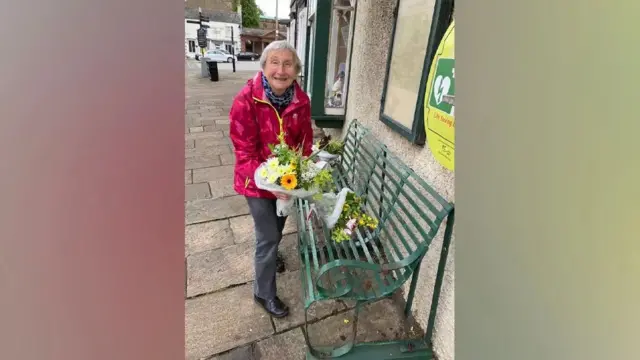 An elderly lady in a pink coat placing a bouquet of yellow and white flowers onto a green bench