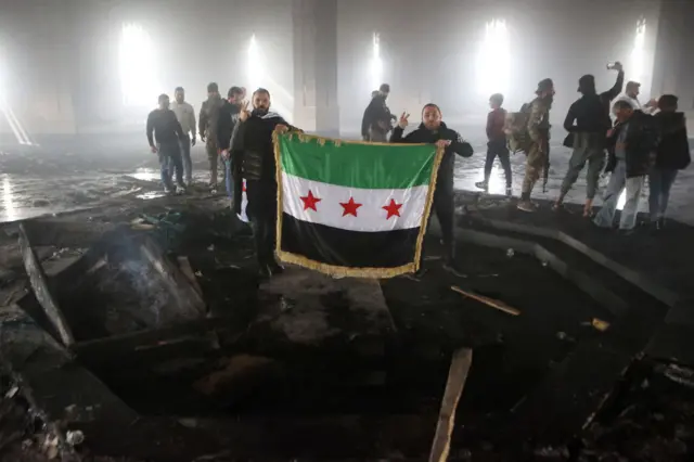 Rebel forces hold up Syria opposition flag at the centre of burned out mausoleum. Parts of charred wood lay on blacked out floor as some men stand taking selfies