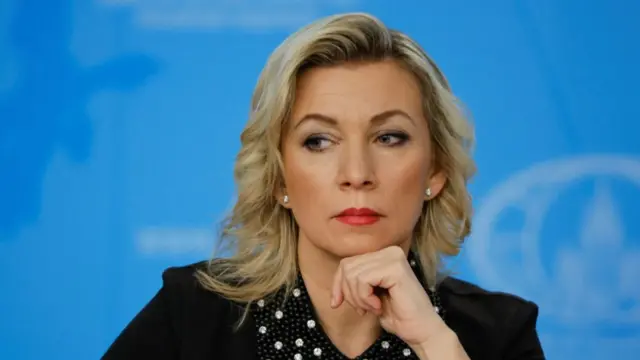 Russian Foreign Ministry spokeswoman Maria Zakharova sitting down, her left hand closed supporting her chin. She's in a black top with decorated collar