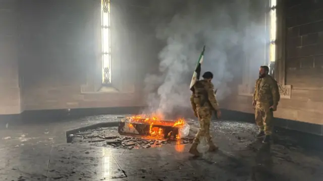 Two men stand next to a smouldering coffin in Qardaha