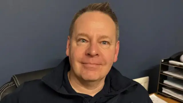 Close up photo of a man's face, he's looking into the camera, wearing a blue fleece jumper with brown hair combed back
