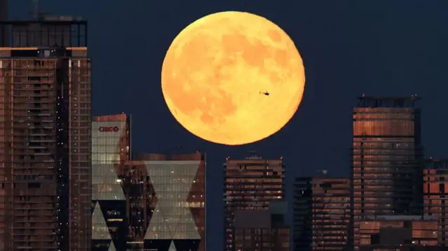 Skyline of Toronto, Canada, with multiple high-story buildings made of glass with a huge yellow moon above them as helicopter flies in the sky
