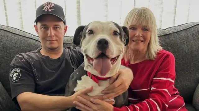 A man and woman sat on a sofa, with a bulldog sat in between them and their arms around the dog, whose mouth is open and tongue sticking out