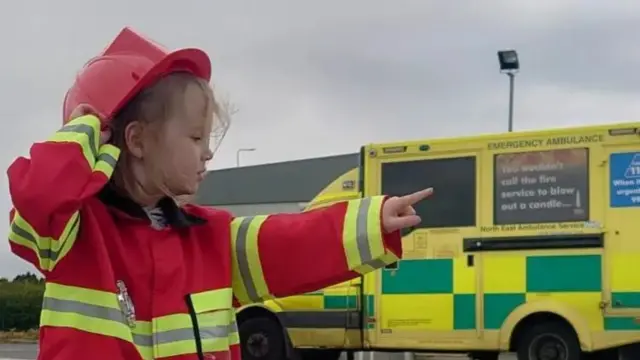 Little girl in red and yellow firefighter uniform and hat stood in front of a yellow and green ambulance