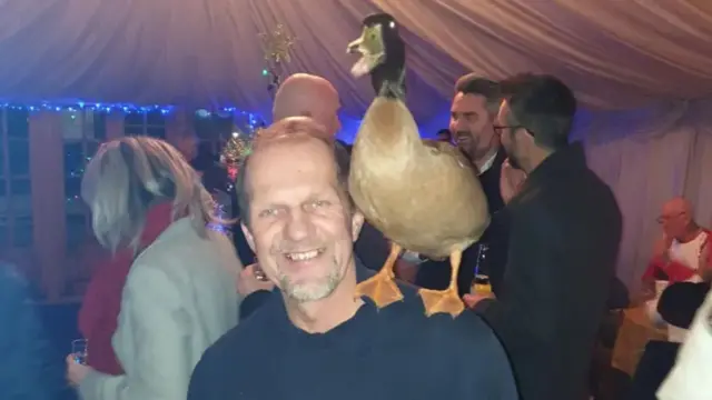 A man stands in a tent with people in the background at a party. He has a duck on his left shoulder, whose beak is partially open