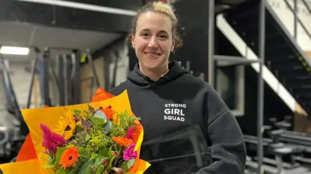 A woman standing in a gym wearing a black jumper that says strong girl squad holding a bunch of flowers with equipment and stairs in the background