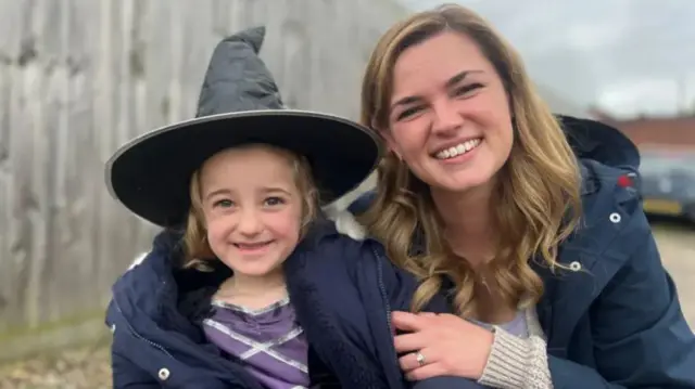 Little girl in a witches hat being held by her mum, sitting next to her on the right, with a driveway and fence behind them