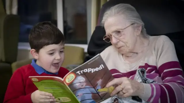 Little boy in red jumper holding one side of a book called trapped as old lady sat next to him in pink jumper holds the other side of the book, they're both reading the same page