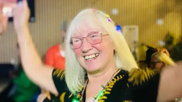 Blonde woman, who is over 65, dances with Christmas lights as a necklace with arms in the air and a smile on her face