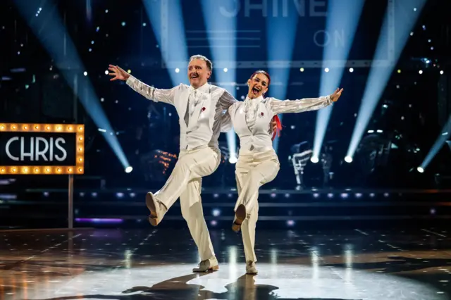 Celebrity dancer Chris McCausland and his professional partner Dianne Buswell - both are wearing white tail coats and are kicking their left legs in the air while holding each other