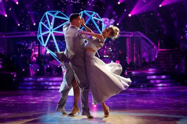 Aljaž Škorjanec wearing a grey tailcoat dances with Tasha Ghouiri who is in white floaty dress decorated with diamante