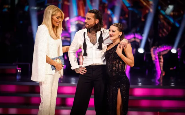 Tess Daly, wearing a white three-piece puts her arm on Pete Wicks' right arm as he stands next to professional dancer Jowita Przystal