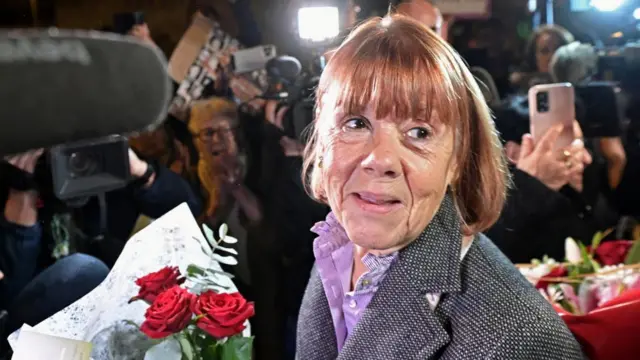 Gisèle Pelicot carries a bunch of flowers, surrounded by the press, film cameras and supporters with placards