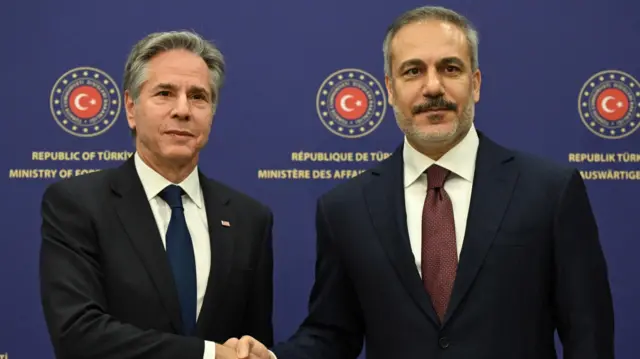 Antony Blinken and Hakan Fidan shaking hands. Both men are wearing black suits, Blinken wearing a dark blue tie while Fidan wears a dark red tie. They're standing in front of a board which reads: 'Republic of Turkiye, Ministry of Foreign Affairs'