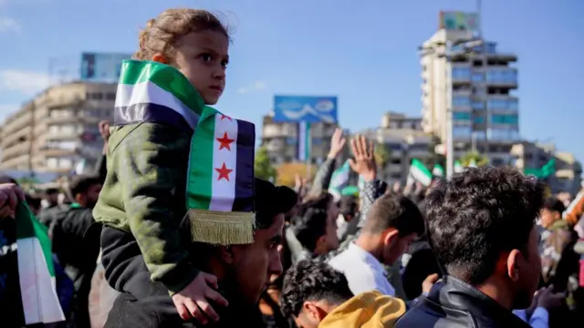 A Syrian man carries a young girl on his shoulders. The child is wearing a green jacket and is wearing the rebel flag